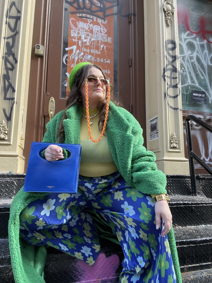 image of kelly-lynne in a green fuzzy coat, green beret, with floral pattern blue and green pants, futuristic glasses with an orange chain, and a blue blocky bag